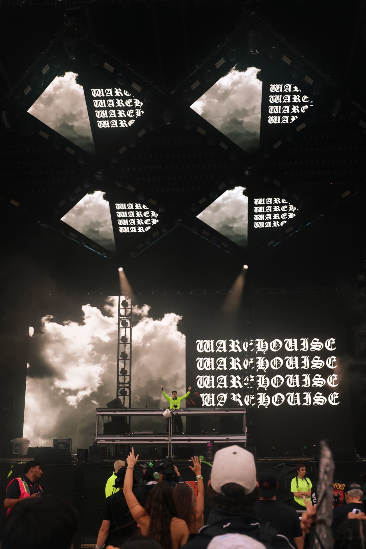 DJ in neon green performing on a warehouse stage with cloud visuals and raised hands