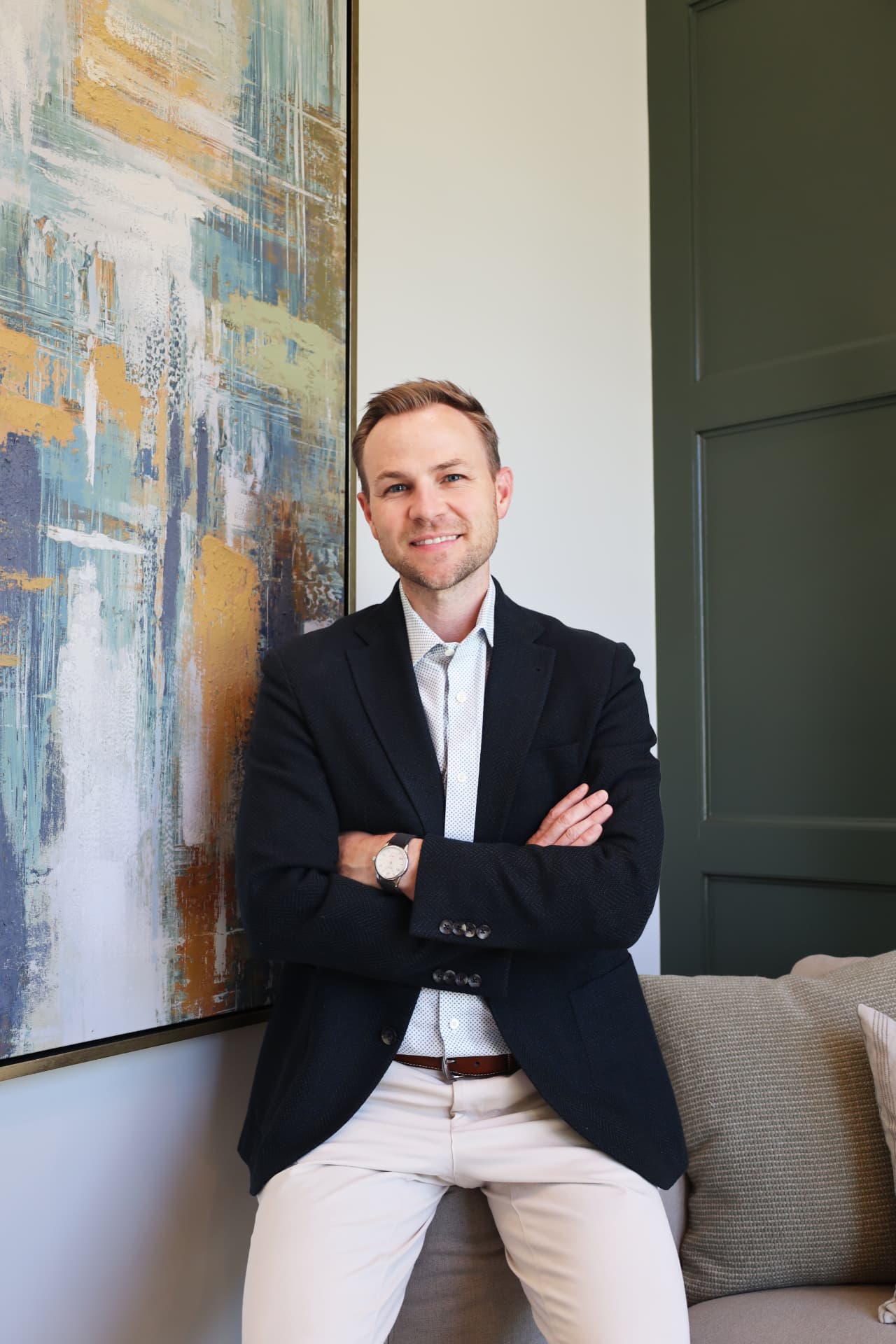 Professional headshot of a man in a navy blazer seated beside abstract art