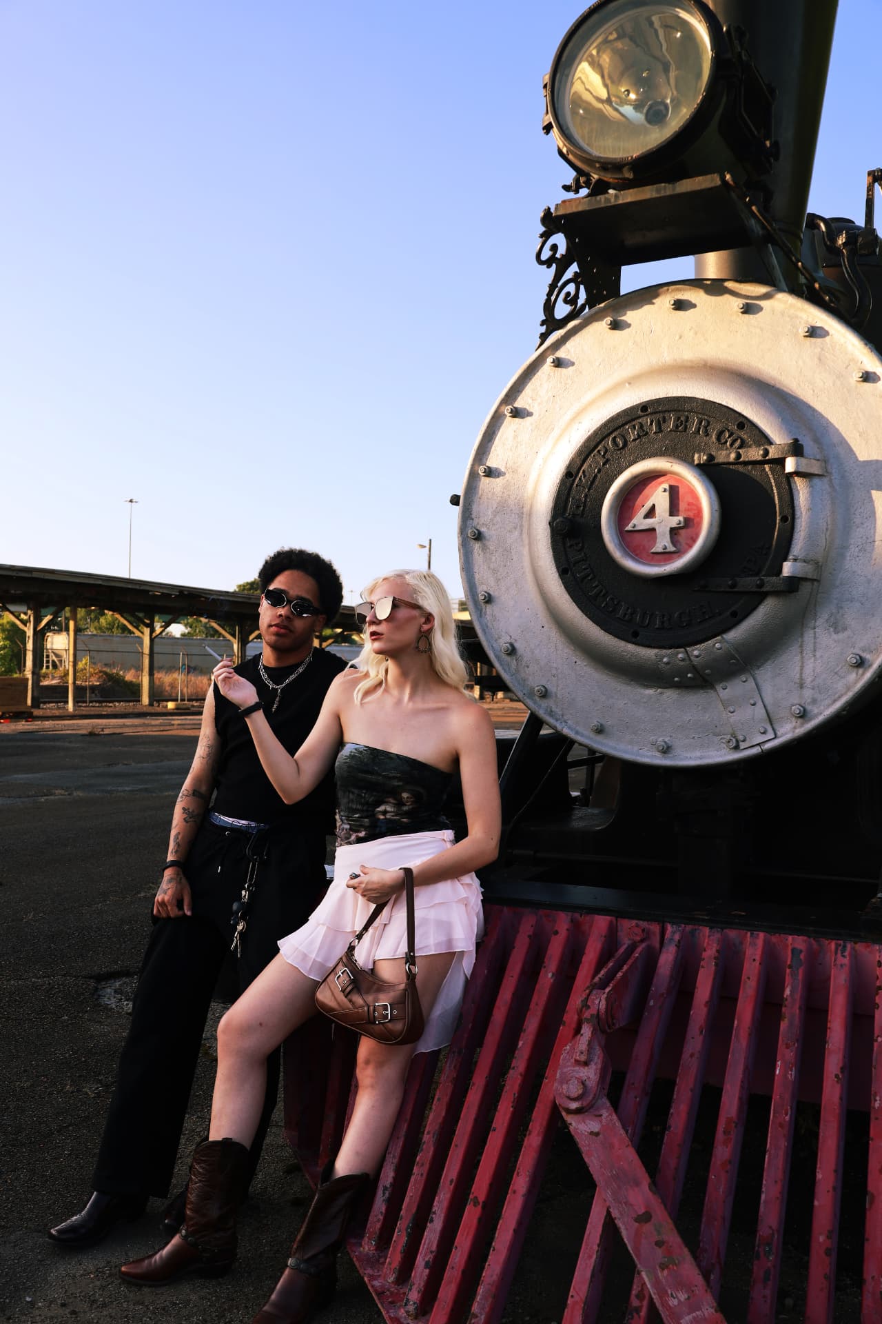 Wide editorial frame of two models leaning on a vintage locomotive under open sky