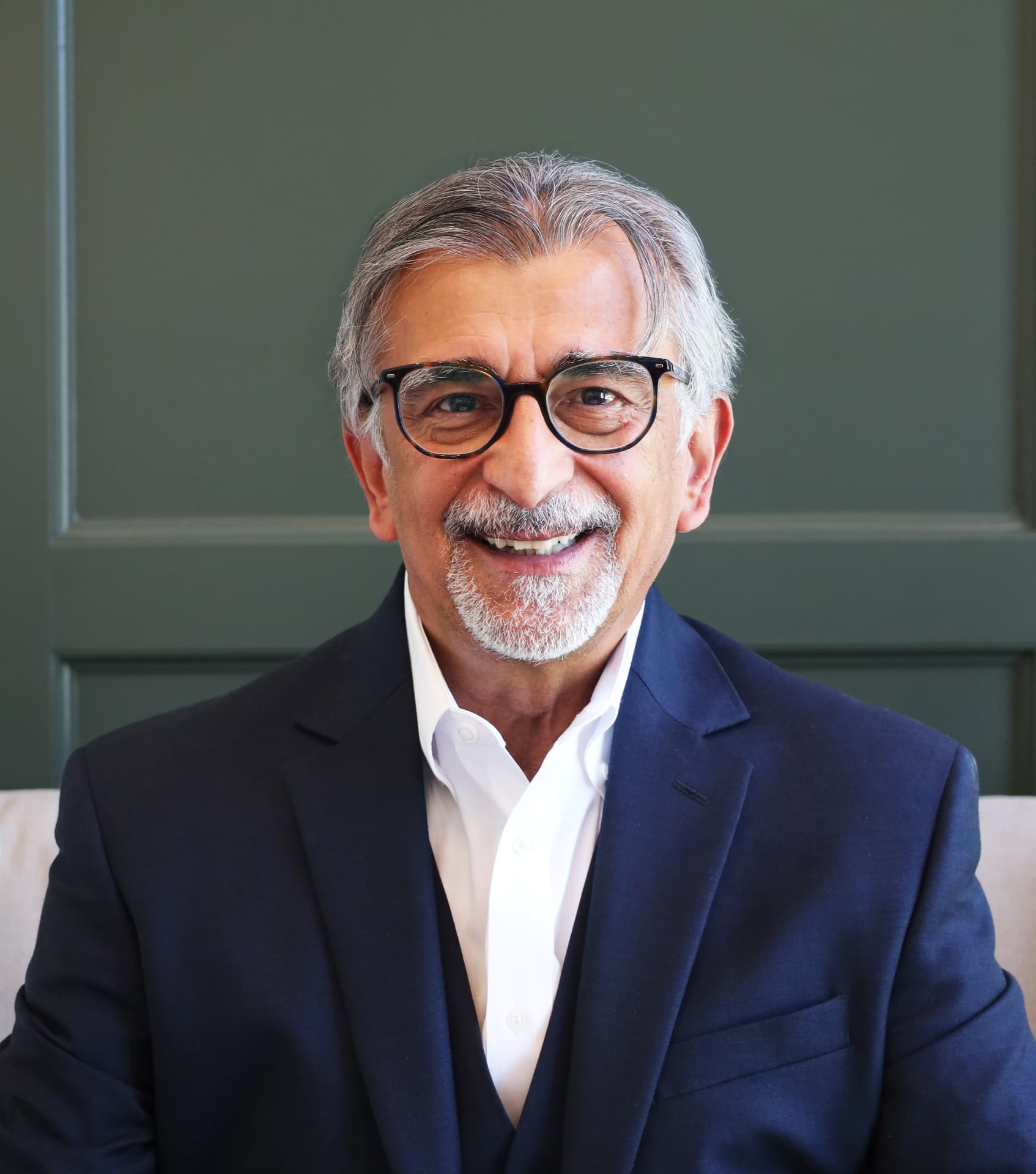 Professional headshot of a man with glasses in a navy suit against a deep green backdrop