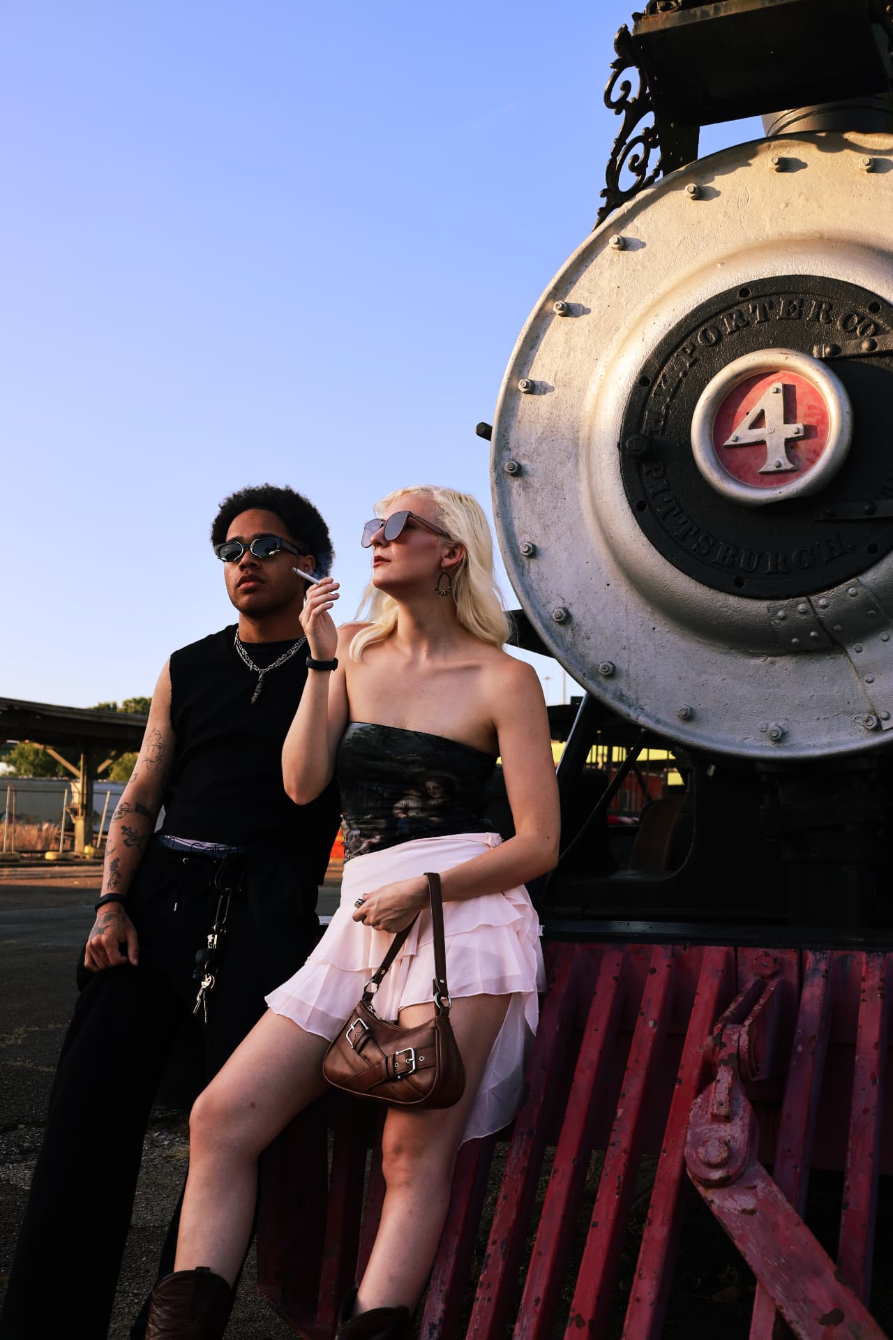 Two models in sunglasses posing against a vintage steam locomotive at golden hour