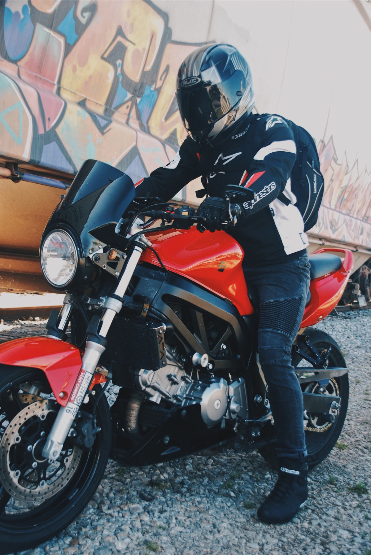 Rider in Alpinestars gear mounting a red naked bike beside a graffiti freight car