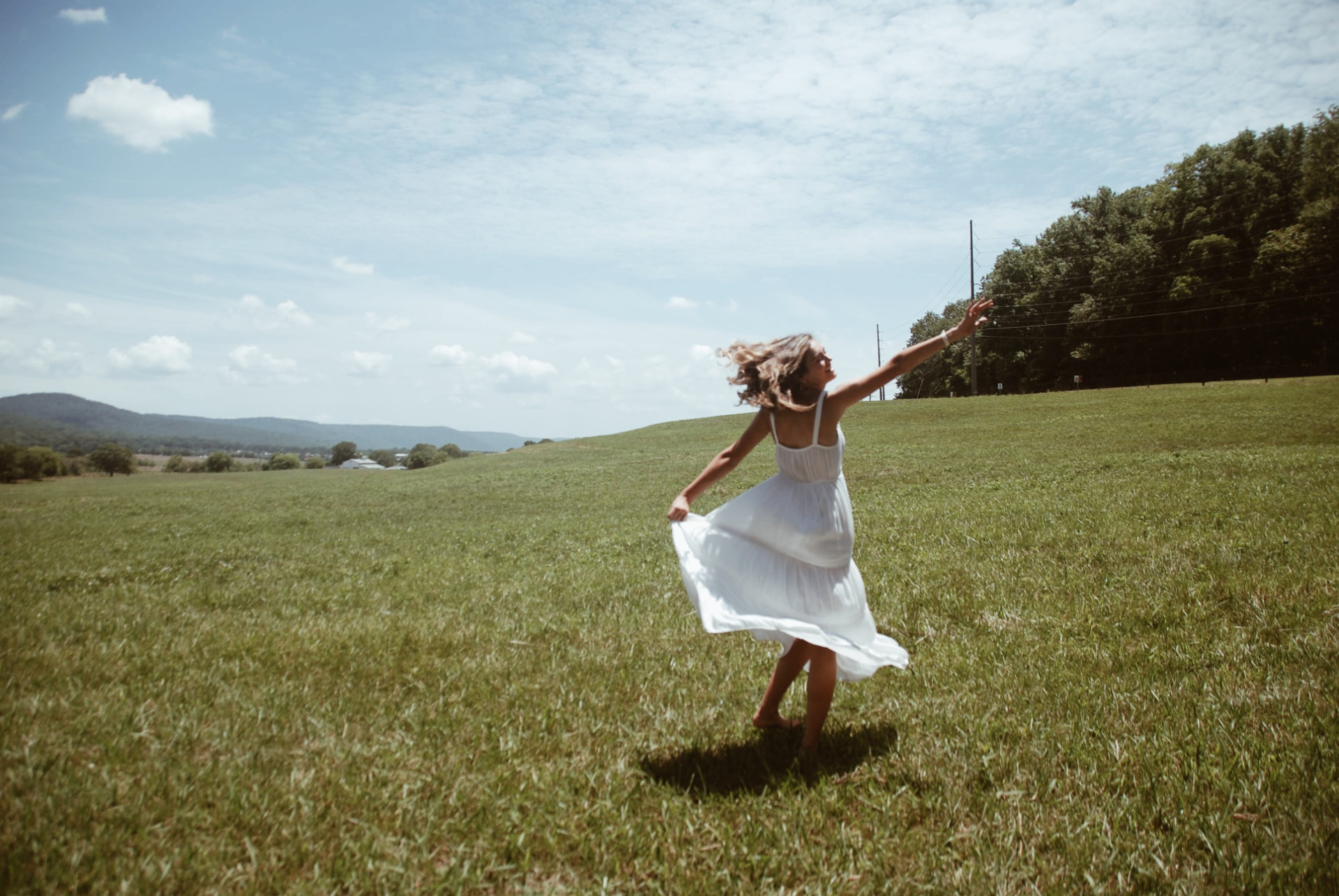 Model in a white dress twirling barefoot across an open field beneath rolling hills