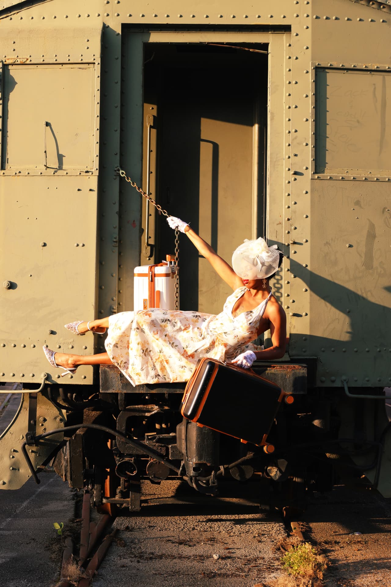 Model reclining with a suitcase against a sunlit vintage rail car