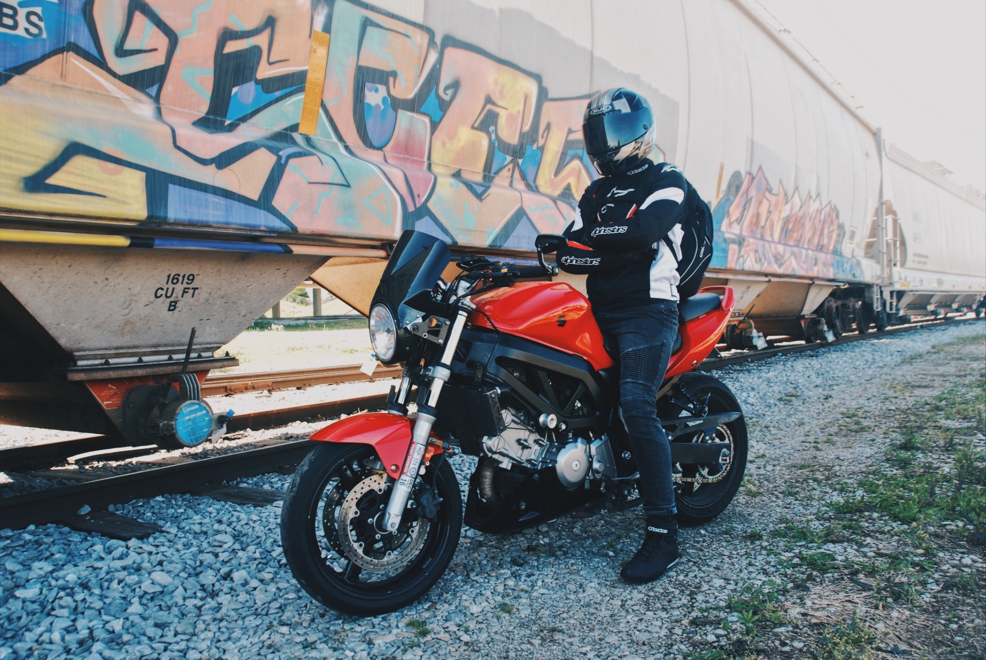 Wide frame of a rider leaning on a red motorcycle along graffiti freight cars