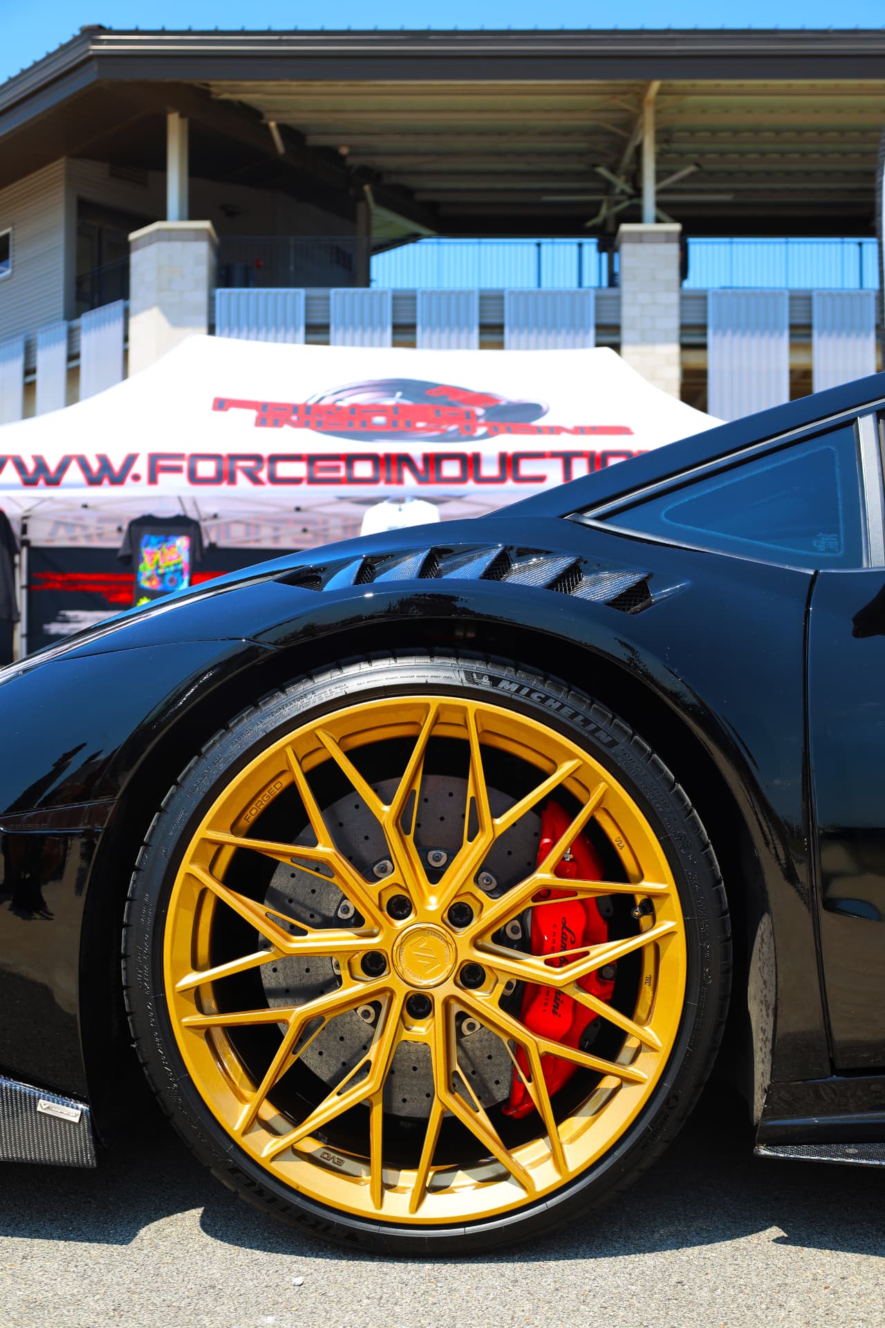 Custom forged gold wheel and red brake caliper detail on a black sports car at an automotive expo