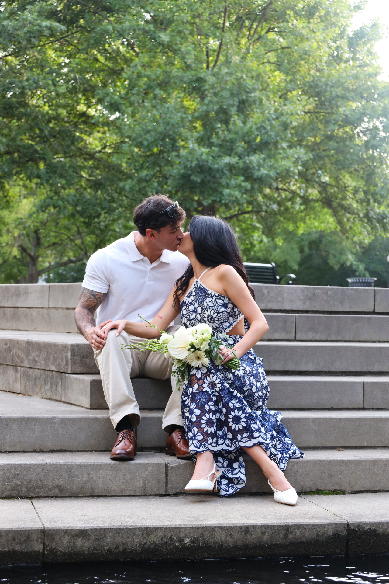 Engaged couple sharing a kiss on stone park steps with bouquet