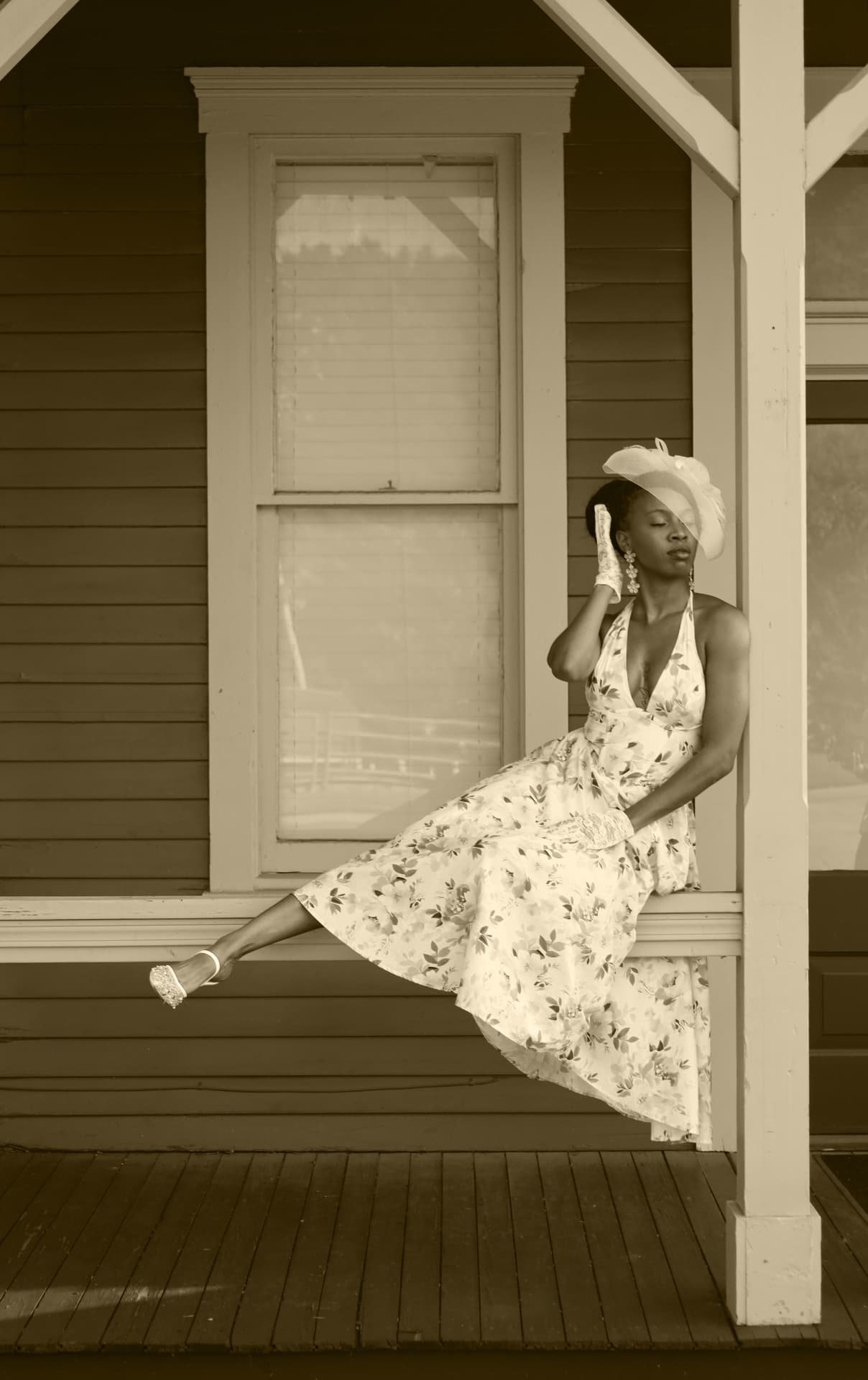 Sepia portrait seated on a porch rail in a floral dress and wide veil hat